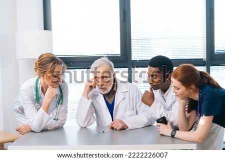Medical team of diverse professional doctors looking at syringe with new coronavirus vaccine on desk and discussing, standing on background of window in hospital with modern light interior.