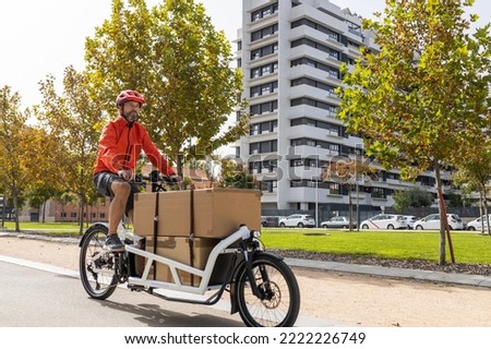 young courier with red clothing and helmet riding cargo bike , riding along the city bike path on his way to deliver a package.