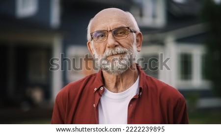 Portrait of a Happy Senior Man with Gray Hair Wearing Glasses and a Red Shirt Standing Outside in Front of a Suburbs Area House. Old Adult Man Posing and Looking at Camera and Smiling. Royalty-Free Stock Photo #2222239895