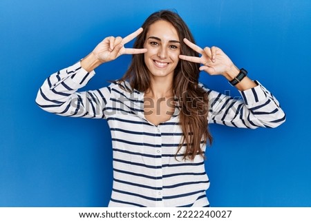 Young hispanic woman standing over blue isolated background doing peace symbol with fingers over face, smiling cheerful showing victory 