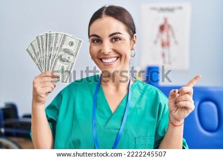 Young hispanic woman holding dollars banknotes working at pain recovery clinic smiling happy pointing with hand and finger to the side 