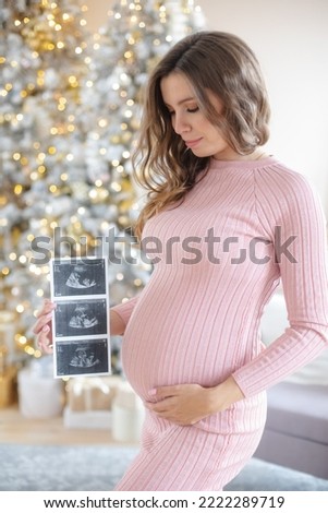 Stylish pregnant woman in pink knitted dress holds an ultrasonic sonogram picture of baby the background of a decorated christmas tree. Prenatal care, motherhood, medicine, pharmaceutics, health care
