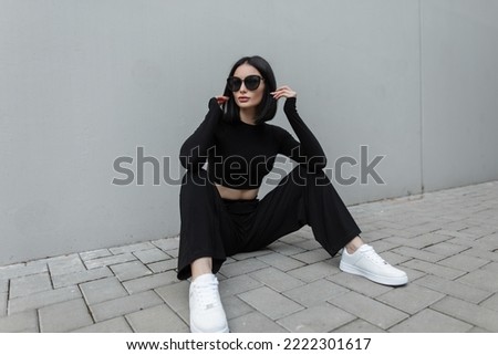 Fashionable young urban woman with black trendy sunglasses in fashion black stylish sports outfit with white sneakers sits on the street near a modern gray building Royalty-Free Stock Photo #2222301617