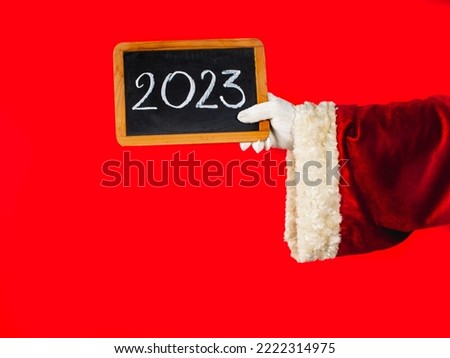 Santa Claus's hand is holding a blackboard with the new year 2023 number written in chalk. Red traditional Santa Claus costume on a background. Handwritting illustration of chalk on black blackboard.