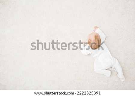 Baby in white bodysuit crawling on knee and arms on light beige home carpet background. Top view. 5 to 6 months old infant development. Empty place for text. Royalty-Free Stock Photo #2222325391