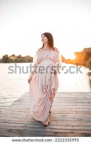 A young pregnant woman in a delicate dress by the river with a beautiful sunset. Beauty, health and gentle motherhood. happy motherhood