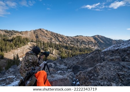 Hunter wearing camo uses his phone to take photos through a spotting scope while deer hunting in the Wyoming mountains