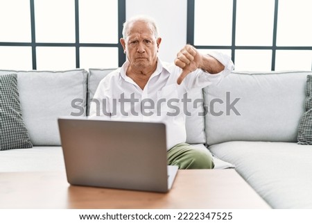 Senior man using laptop at home sitting on the sofa looking unhappy and angry showing rejection and negative with thumbs down gesture. bad expression. 