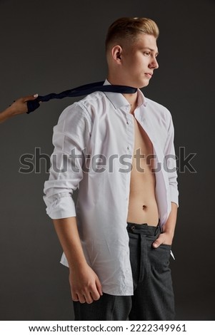Portrait of young man in white shirt posing with serious expression isolated over grey background. Woman pulling tie. Concept of men's health, natural beauty, body and skin care.