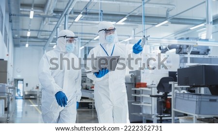 Two Scientists Walking in a Heavy Industry Factory in Sterile Coveralls and Face Masks, Using Laptop Computer. Examining Industrial Machine Settings and Configuring Production Functionality. Royalty-Free Stock Photo #2222352731