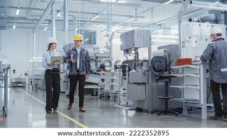 Wide Shot: Two Heavy Industry Employees in Hard Hats Discussing Job Assignments at the Factory, Using Laptop Computer. Asian Engineer and Technician at Work Smiling Royalty-Free Stock Photo #2222352895