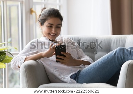 Pretty Indian woman sit on comfortable armchair spend weekend at home with smartphone, making call or order, have fun on internet looks satisfied buying e-services use electronic commerce application