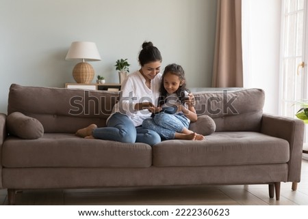 Indian woman and little daughter spend time using digital tablet, sit together on couch at home, buying goods, purchasing toys through retail webstore enjoy new application for fun or kid development Royalty-Free Stock Photo #2222360623