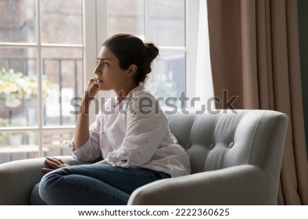 Sad upset young Indian woman thinking while sit on armchair and staring out window. Personal troubles and break up, goes through difficult life situation, search solution, ponders seated alone at home Royalty-Free Stock Photo #2222360625