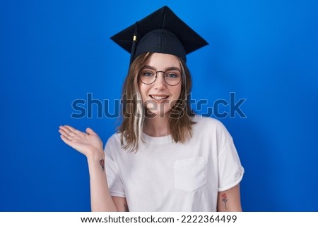 Blonde caucasian woman wearing graduation cap smiling cheerful presenting and pointing with palm of hand looking at the camera. 