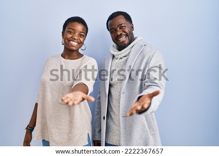 Young african american couple standing over blue background together smiling cheerful offering palm hand giving assistance and acceptance. 