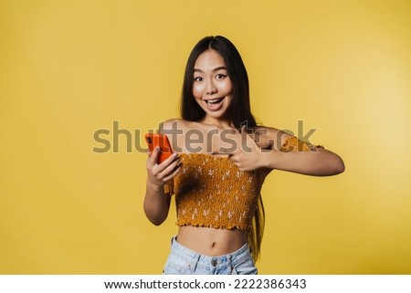 Young asian woman smiling and pointing finger at her cellphone isolated over yellow background
