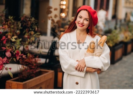 Portrait of a pretty woman in a red beret and a white cardigan with baguettes in her hands strolling through the autumn city. Royalty-Free Stock Photo #2222393617