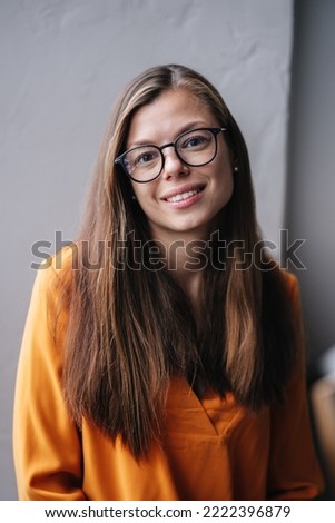 Vertical shoot of cute brunette young hispanic woman in glasses, orange blouse sitting on windowsill looks at camera toothy smiling against grey wall indoors. Satisfied housewife relaxing at home.
