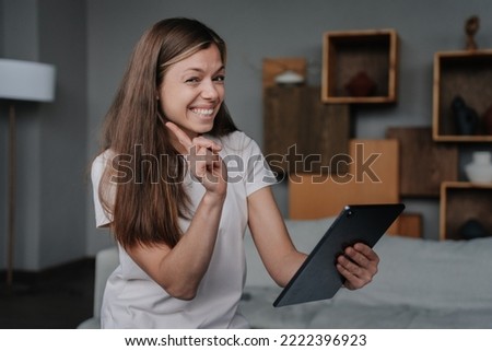 Cheerful young brunette American woman holding tablet smiles wide, warns shakes index finger looking at camera, sitting at home. Adorable housewife asks to not be naughty. Communication and leisure. Royalty-Free Stock Photo #2222396923