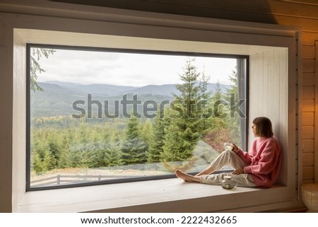 Woman sits with phone on window sill and enjoys scenic view on mountains while resting in wooden house on nature. Recreation and escaping to nature concept Royalty-Free Stock Photo #2222432665