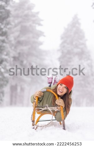 Beautiful young woman lying on a sledge having fun sliding down the hill in the snow while spending winter vacation in the mountains