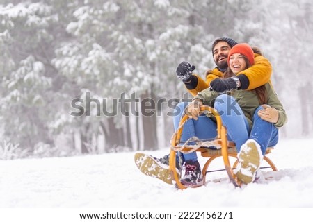 Beautiful young couple in love having fun spending winter vacation in mountains, sitting and hugging on sled, sliding down the hill in the snow