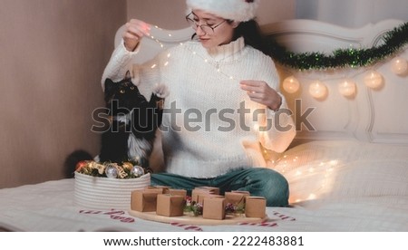Beautiful caucasian teenage girl in santa claus hat plays with purebred cat with burning garland while sitting on bed with advent calendar and christmas decorations in bedroom, closeup side view.