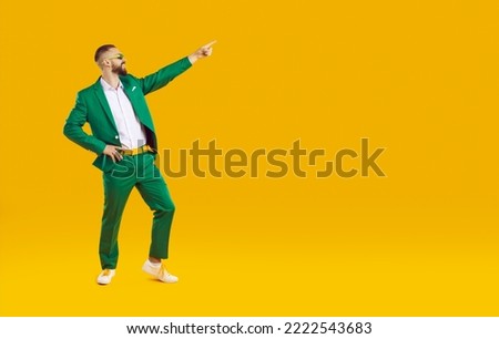 Confident handsome showman in green suit, white shirt and sunglasses showing pointing index finger on the right on yellow background, full length portrait. Banner for advertisement, marketing.