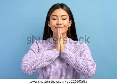 Gorgeous woman keeps hands in yoga gesture, has calm facial expression, keeping palms pressed together and closed eyes. Indoor studio shot isolated on blue background 
