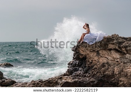 A woman in a storm sits on a stone in the sea. Dressed in a white long dress, waves crash against the rocks and white spray rises above her.