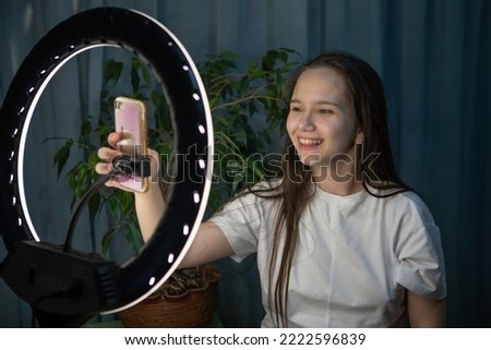 Teenage girl, blogger, smiling at the camera for a social network using a smartphone and a ring lamp in the children's room. Teen fooling around in front of the camera. Selective focus