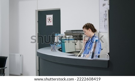 Woman nurse answering landline phone call at reception desk in healthcare facility, using cord telephone line to make medical appointments. Working at hospital registration counter. Handheld shot.