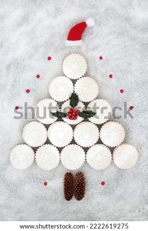 Surreal Christmas tree mince pies concept shape,with santa hat decoration, pine cones and winter holly with red berries on snow background. Festive food design for Xmas and New Year.
