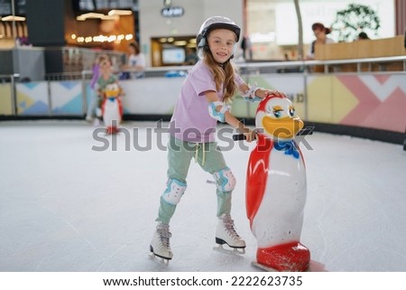Girl trains figure skating on ice. Support for learning to skate in the form of a penguin. Protective ammunition helmet and elbow pads. Selective focus, blurred background.