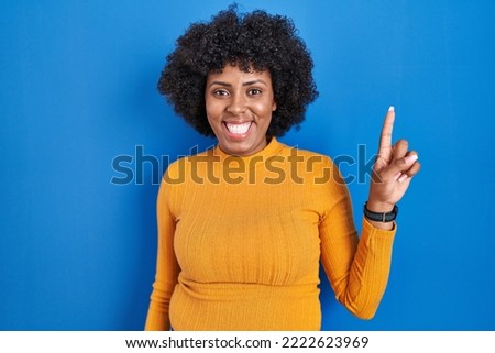 Black woman with curly hair standing over blue background showing and pointing up with finger number one while smiling confident and happy. 