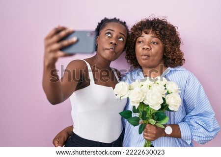 Two african women taking a selfie photo with flowers making fish face with mouth and squinting eyes, crazy and comical. 