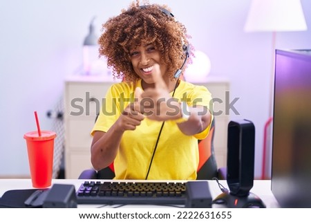 Young hispanic woman with curly hair playing video games wearing headphones approving doing positive gesture with hand, thumbs up smiling and happy for success. winner gesture. 