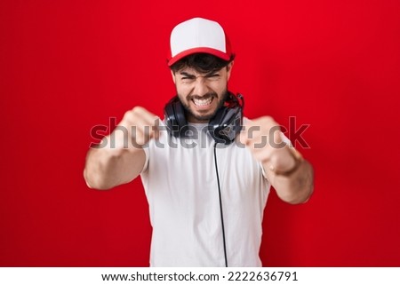 Hispanic man with beard wearing gamer hat and headphones angry and mad raising fists frustrated and furious while shouting with anger. rage and aggressive concept. 