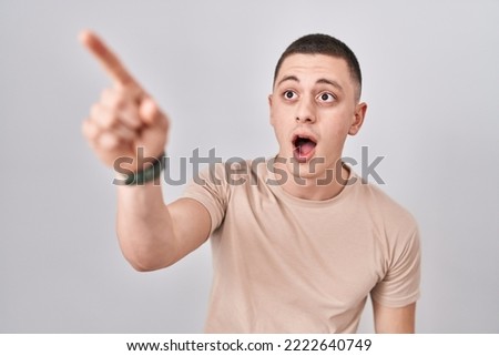 Young man standing over isolated background pointing with finger surprised ahead, open mouth amazed expression, something on the front 