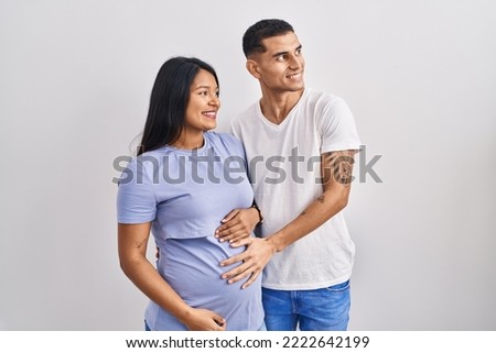 Young hispanic couple expecting a baby standing over background looking away to side with smile on face, natural expression. laughing confident. 