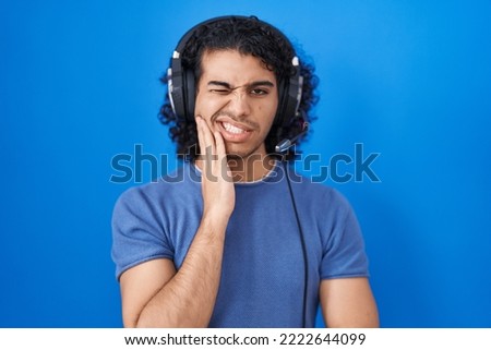 Hispanic man with curly hair listening to music using headphones touching mouth with hand with painful expression because of toothache or dental illness on teeth. dentist 