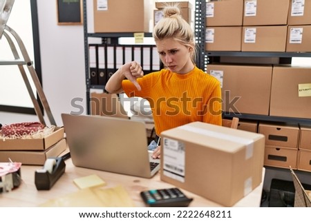 Young blonde woman working at small business ecommerce using laptop with angry face, negative sign showing dislike with thumbs down, rejection concept 