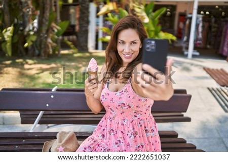 Brunette woman enjoying a summer day sitting on a bench at the park eating a ice cream cone taking a selfie picture