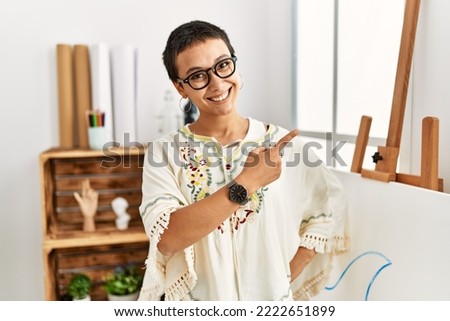 Young hispanic woman with short hair at art studio cheerful with a smile of face pointing with hand and finger up to the side with happy and natural expression on face 