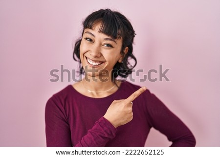 Young beautiful woman standing over pink background cheerful with a smile of face pointing with hand and finger up to the side with happy and natural expression on face 