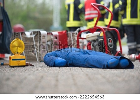 A heavy training dummy lies on the ground after the fire brigade exercise, in the background there is various equipment and firefighters are discussing it