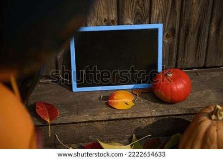 A black black chalkboard, with copy advertising space for text, a bright orange pumpkin and fallen dry leaves on the doorstep, against a wooden background. Autumn. Halloween. Thanksgiving concept