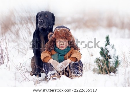                      A little boy in winter clothes is sitting on the snow with a dog (black labrador retriever) looking at the camera. Winter walk.  Human and dog friendship concept. 