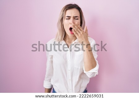 Young beautiful woman standing over pink background bored yawning tired covering mouth with hand. restless and sleepiness. 